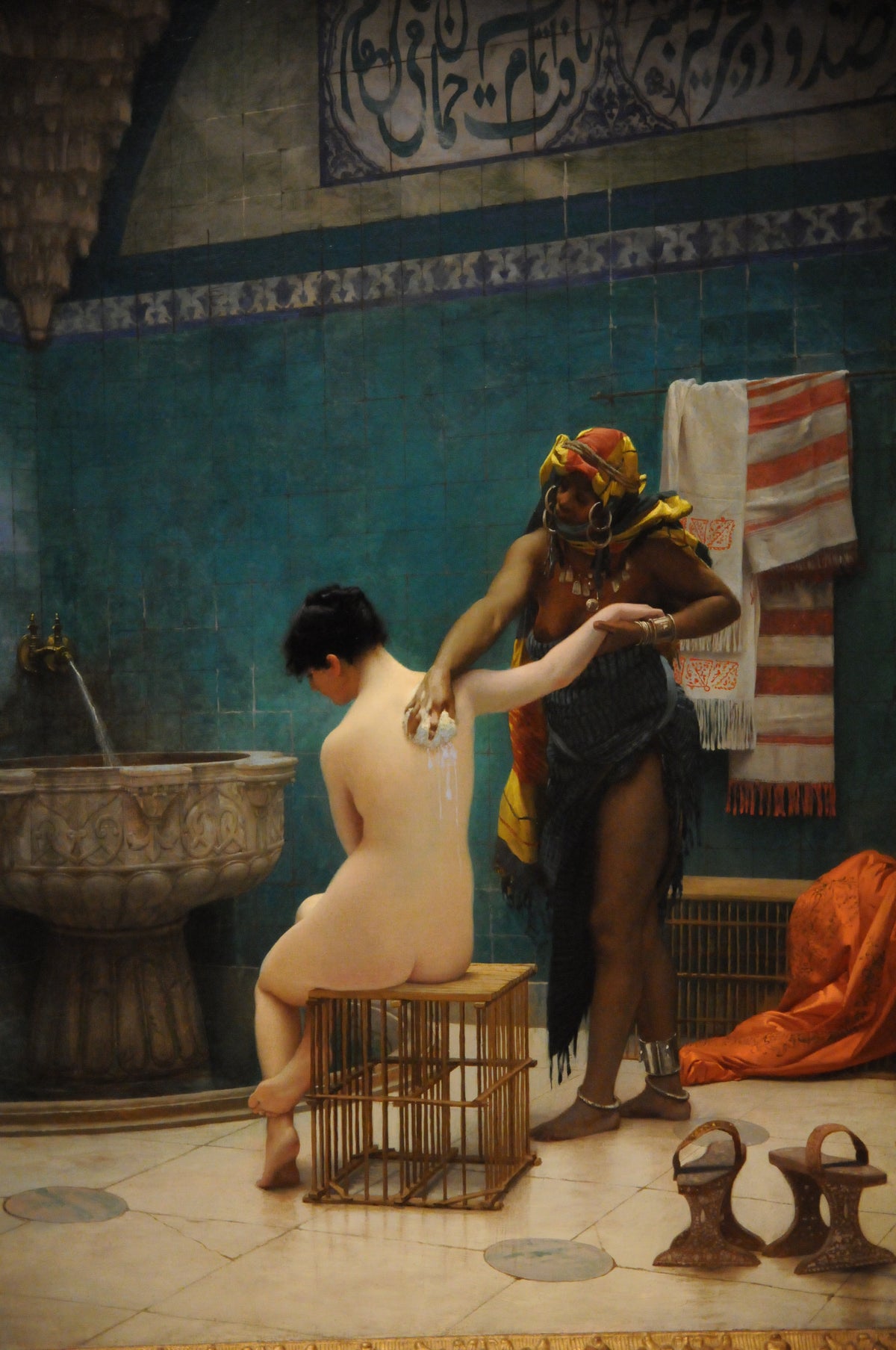 A bath attendant washes a seated woman's back beside a carved stone basin, surrounded by turquoise tiles and patterned textiles.