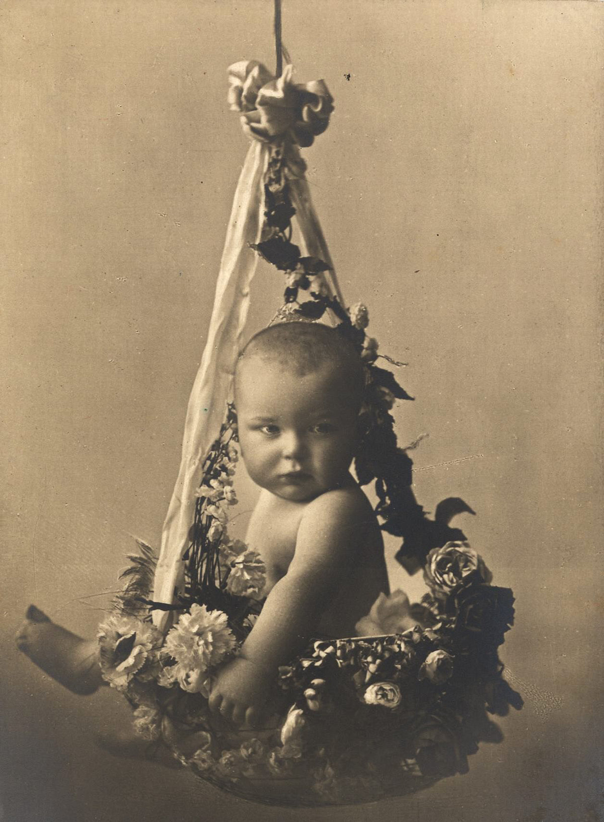 A baby sits in a flower-covered hanging basket in a softly shaded sepia portrait with trailing ribbons and foliage.