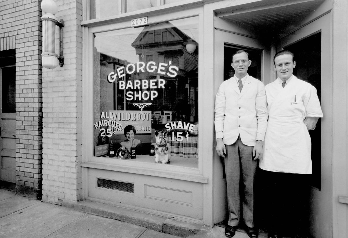 Two men stand in the doorway of George's Barber Shop beside a window filled with period lettering and haircut prices, captured in black and white.