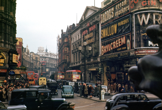Red double-decker buses and dark motorcars fill a busy London street beneath large period advertising signs and a statue in the foreground.